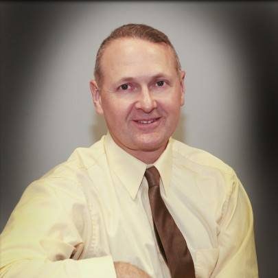 A man with short, light brown hair, wearing a pale yellow shirt and brown tie, sits and smiles at the camera against a neutral, shaded background.