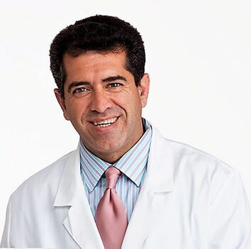 A man with short dark hair, wearing a white lab coat over a striped shirt and pink tie, smiles at the camera against a plain white background.