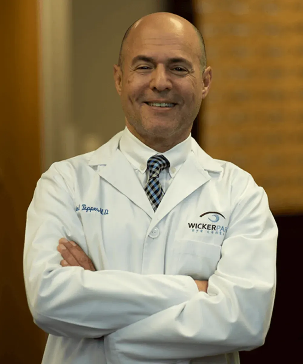 A smiling man in a white lab coat with Wickepan Eye Center on it stands with his arms crossed. He is wearing a striped tie and has a bald head. The background is softly blurred.