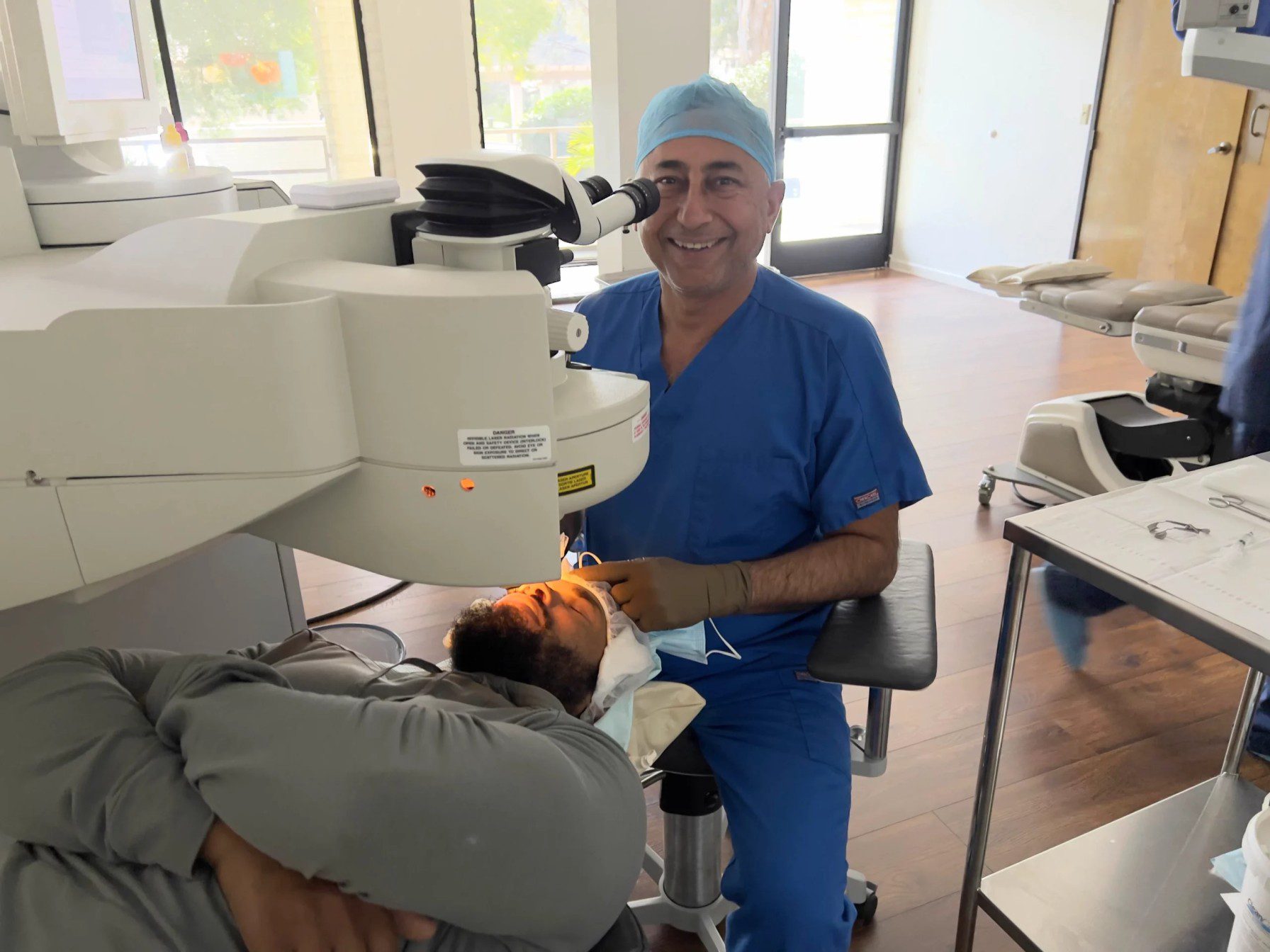 A doctor in blue scrubs and a surgical cap smiles while performing an eye procedure using a large microscope on a patient lying down in a medical clinic.
